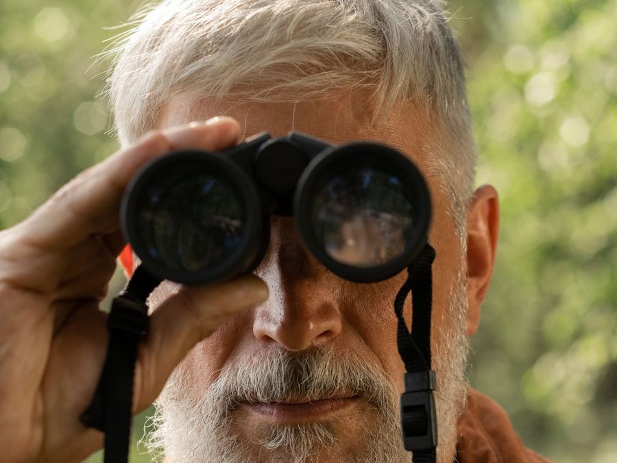An older man looks through binoculars in a sunlit forest