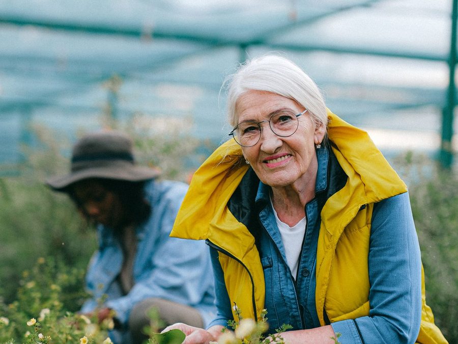 An older woman smiles among the flowers in a bright greenhouse