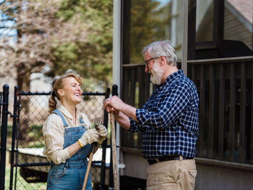A laughing woman and an older man chat over a garden fence on a sunny afternoon
